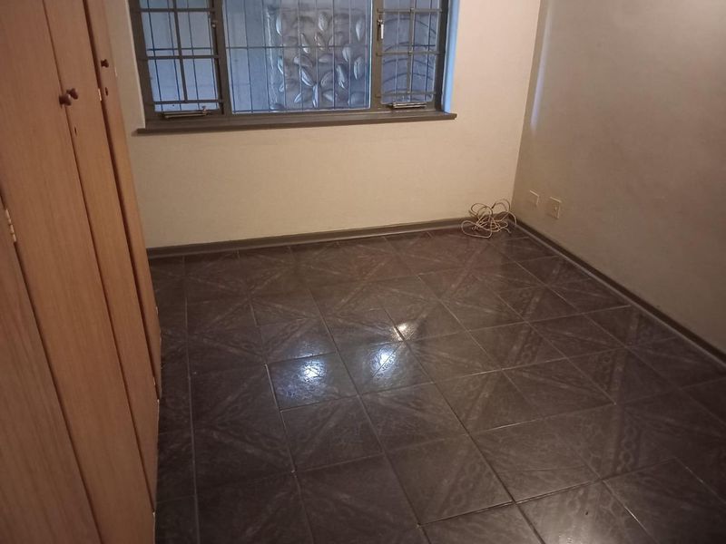 An empty bedroom featuring dark tiled flooring, built-in wooden cupboards, and a window fitted with security bars.
