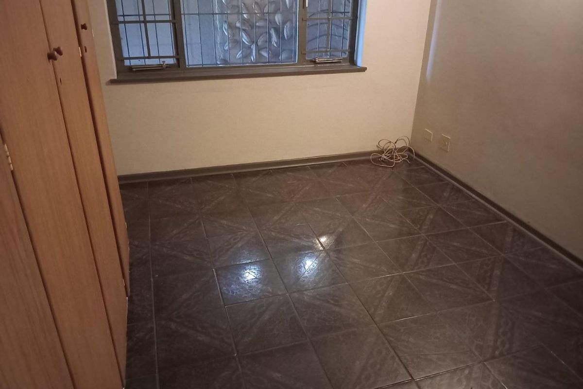 An empty bedroom featuring dark tiled flooring, built-in wooden cupboards, and a window fitted with security bars.