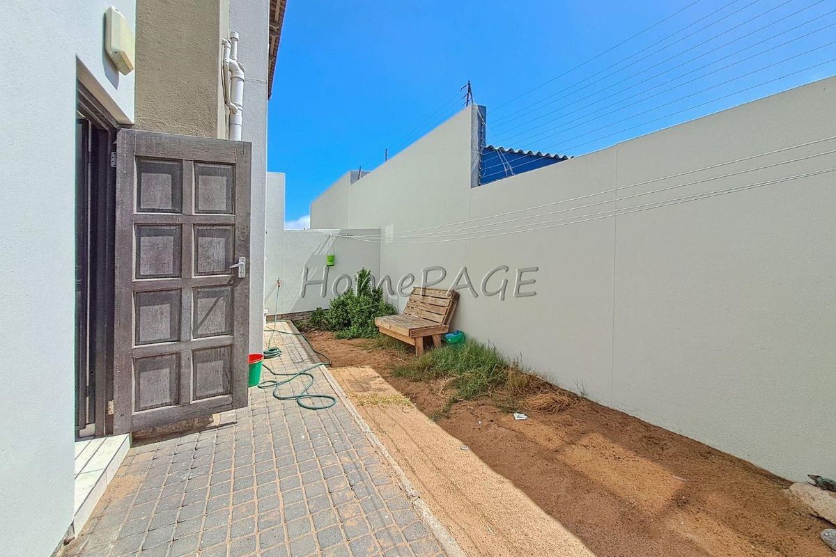 A rear exterior view of the townhouse featuring a paved walkway, a high perimeter wall with electric fencing, and an open garden space.