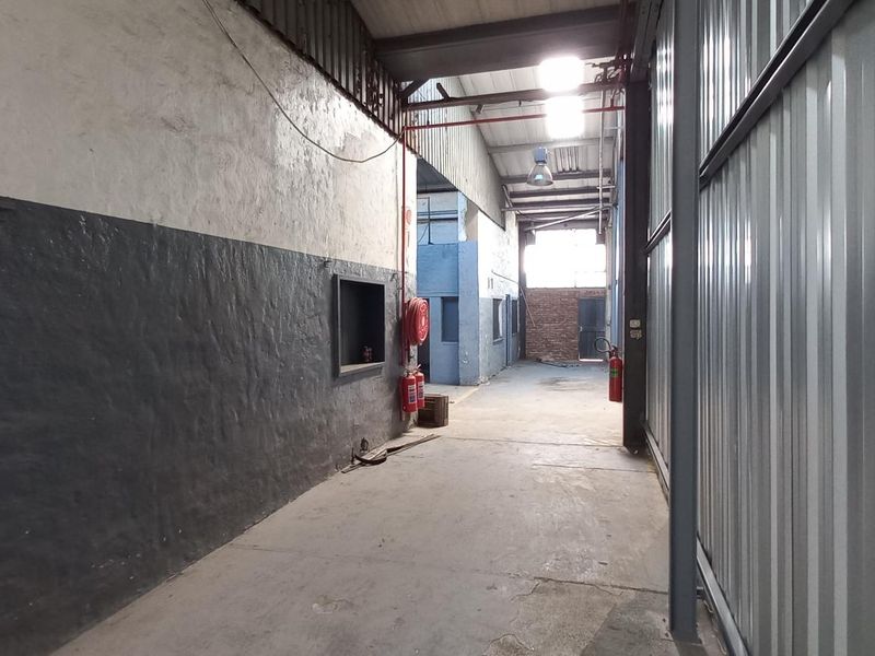 An industrial utility corridor featuring concrete flooring, exposed steel roof structure, and corrugated metal wall cladding, equipped with fire safety equipment.