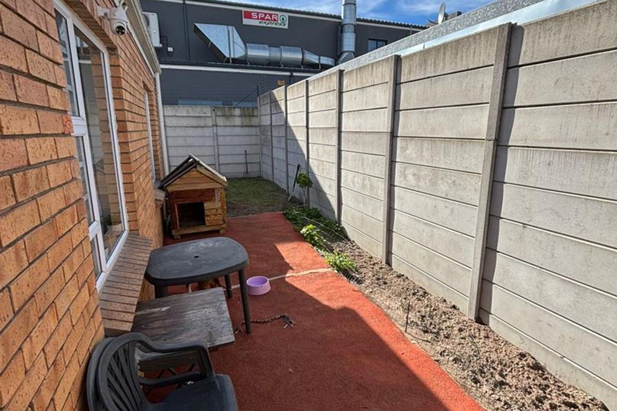 A narrow rear exterior space featuring a brick facade, paved patio area, and a high concrete boundary wall adjacent to commercial property.