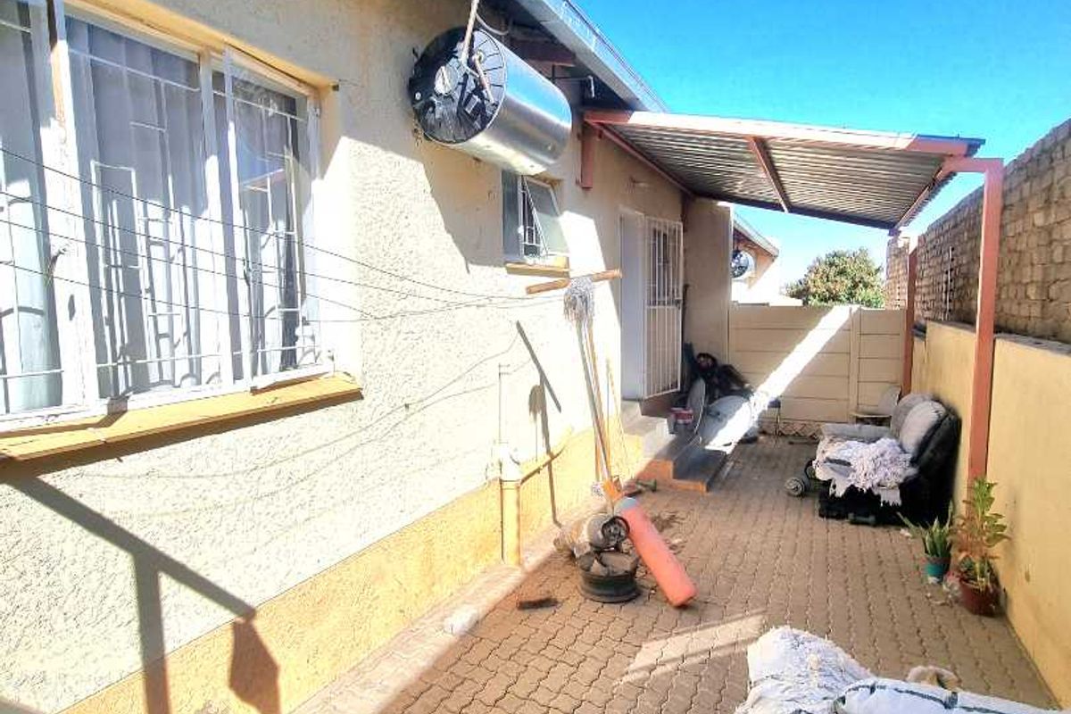 Rear exterior area featuring a paved patio, metal awning, and external geyser unit, enclosed by a boundary wall.