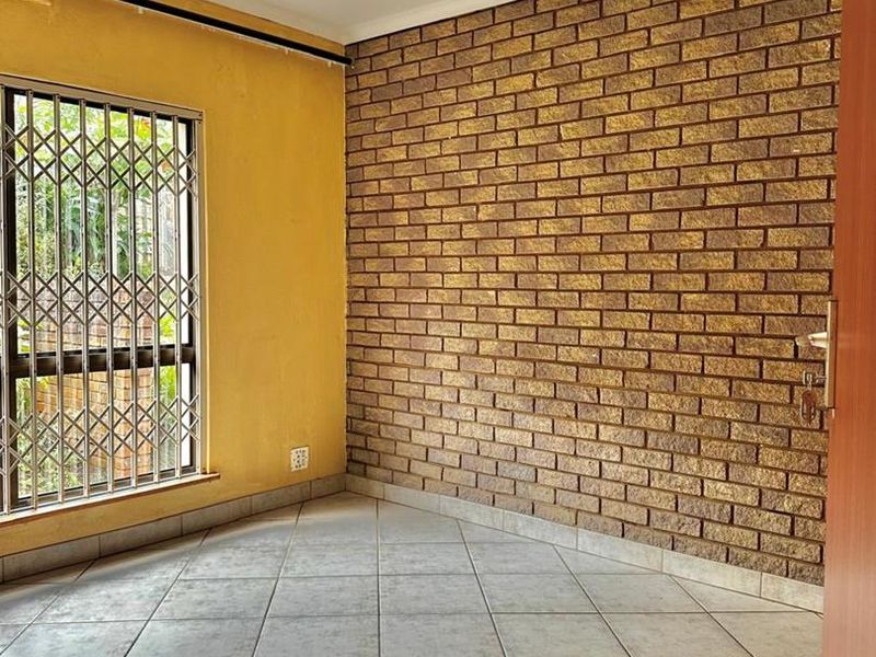 An empty bedroom featuring a prominent exposed brick wall, neutral ceramic floor tiles, and a secure window with a metal security gate.