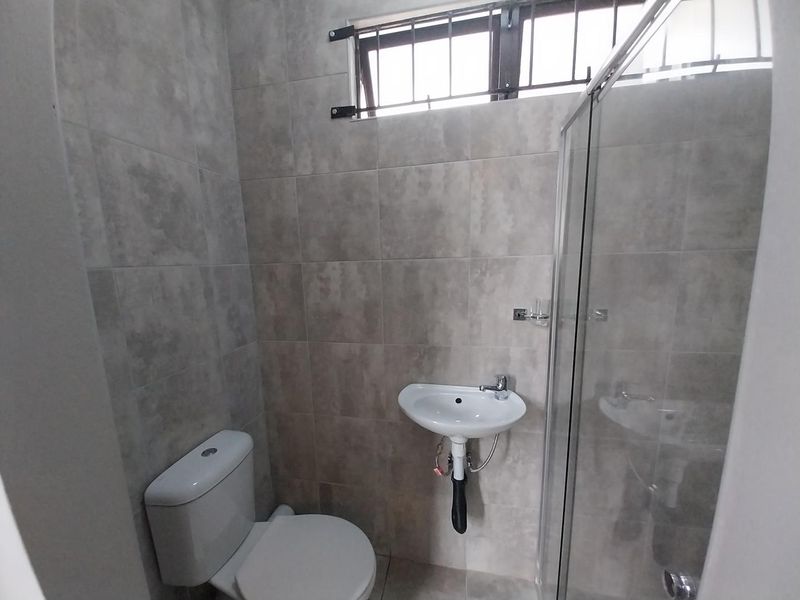 A modern, compact bathroom featuring neutral grey wall tiles, a wall-mounted basin, a toilet, and a glass-enclosed shower.