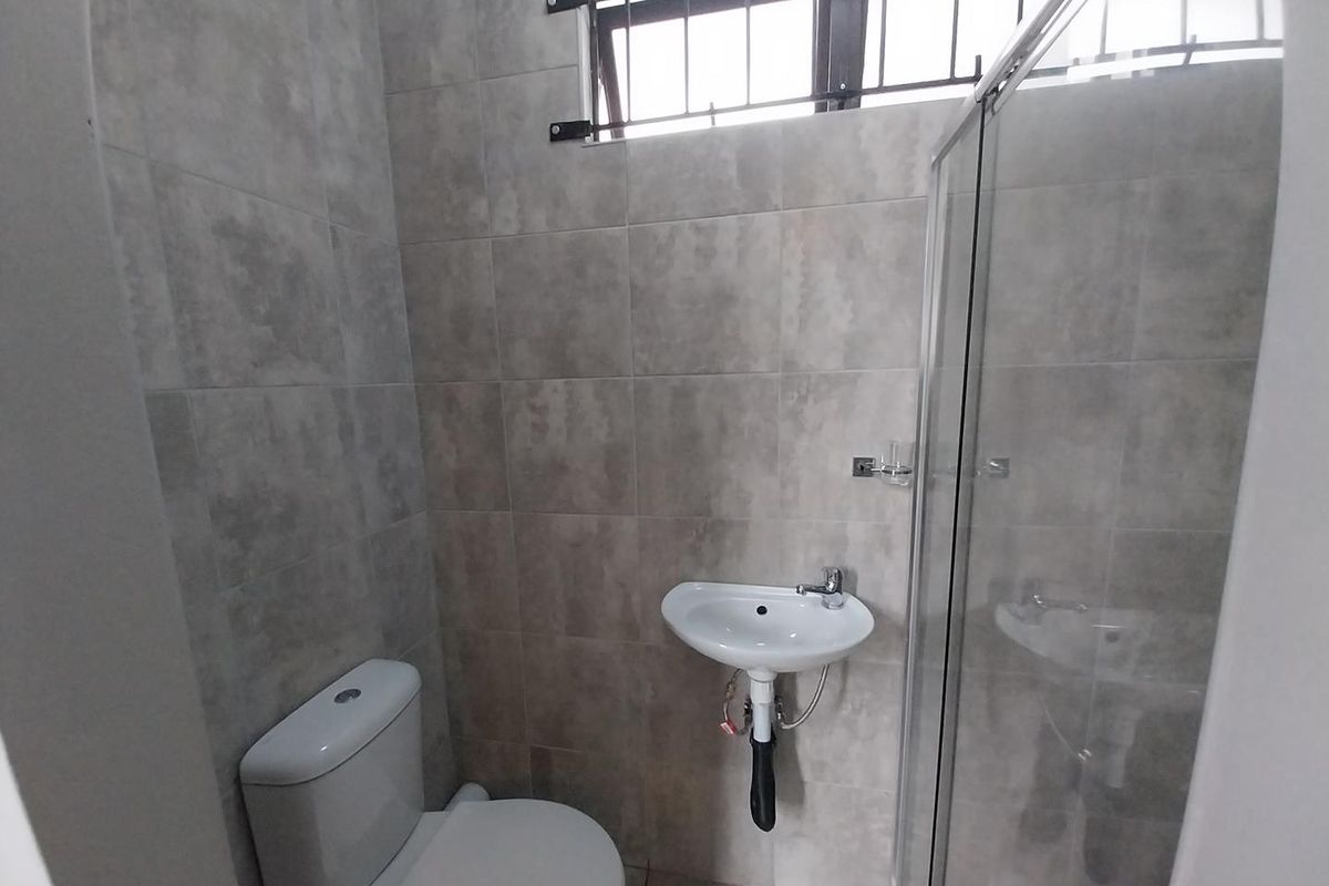 A modern, compact bathroom featuring neutral grey wall tiles, a wall-mounted basin, a toilet, and a glass-enclosed shower.