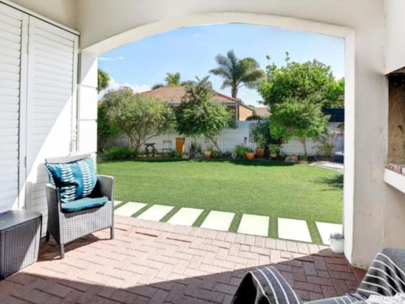 Covered patio featuring brick paving, a built-in braai, and shutter doors, overlooking a well-maintained garden with a lawn and paved stepping stones.