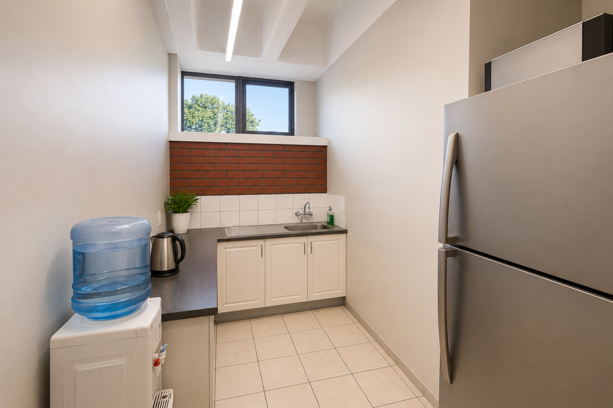 A compact kitchenette area featuring built-in cabinetry, a stainless steel sink, tiled splashback, and a brick accent wall under a high ceiling.