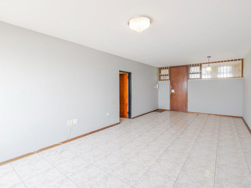 An empty, open-plan living area featuring neutral tiled flooring, a timber entrance door with security gate, and a flush mount ceiling light fixture.