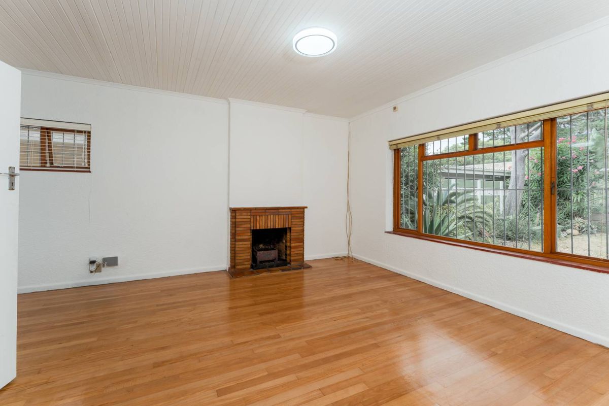 A bright, empty living room featuring polished timber flooring, a classic brick fireplace, and large windows with security bars overlooking the garden.