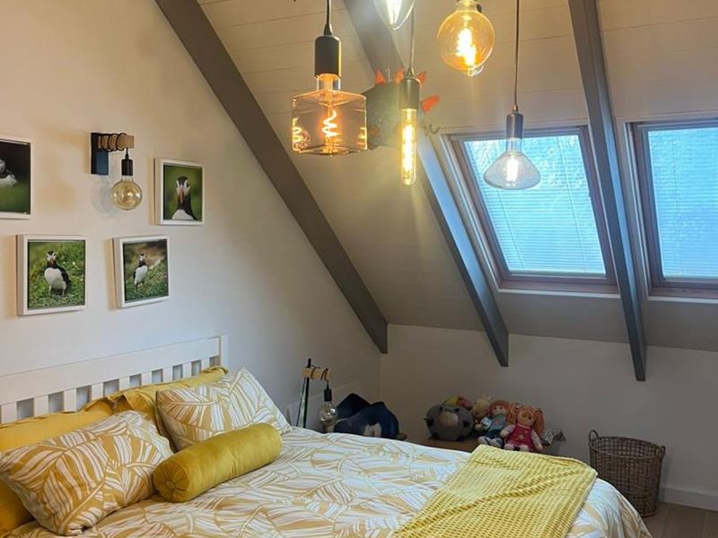 A cozy attic bedroom featuring exposed timber beams, skylight windows, and modern pendant lighting, creating a warm and inviting atmosphere.