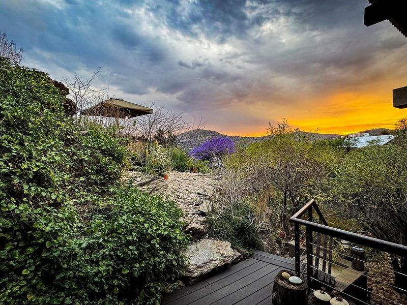 Scenic elevated view overlooking a landscaped garden and natural hillside terrain at sunset, featuring a wooden deck and metal railing.