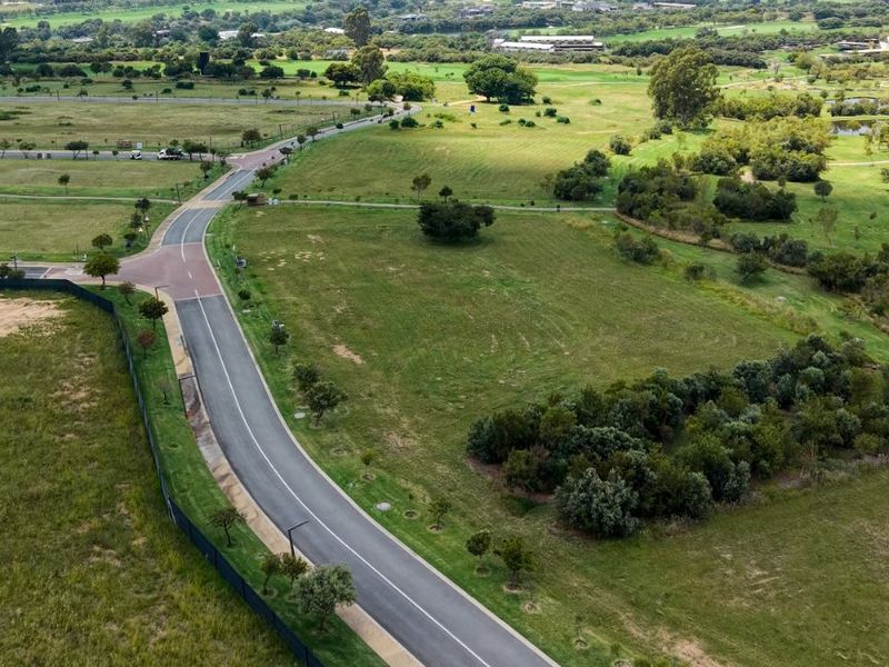 An aerial view of a vacant residential plot featuring established road infrastructure, perimeter fencing, and surrounding green landscape.