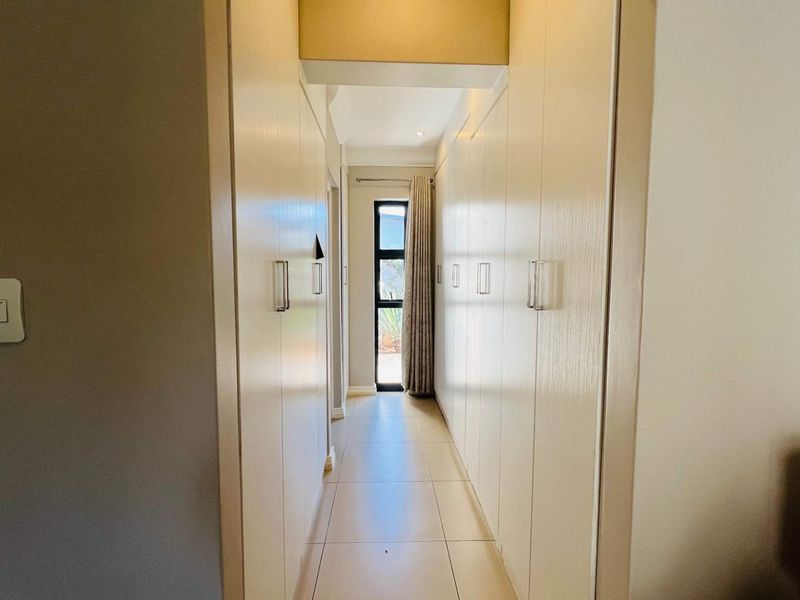 A bright hallway featuring extensive built-in storage cupboards and neutral tiled flooring, leading towards a windowed exit.