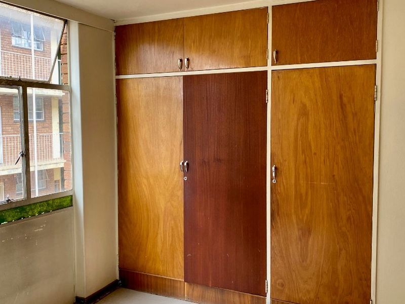 A vacant bedroom featuring original built-in wooden cupboards, a metal-framed window, and basic ceiling lighting, offering a blank canvas for renovation.