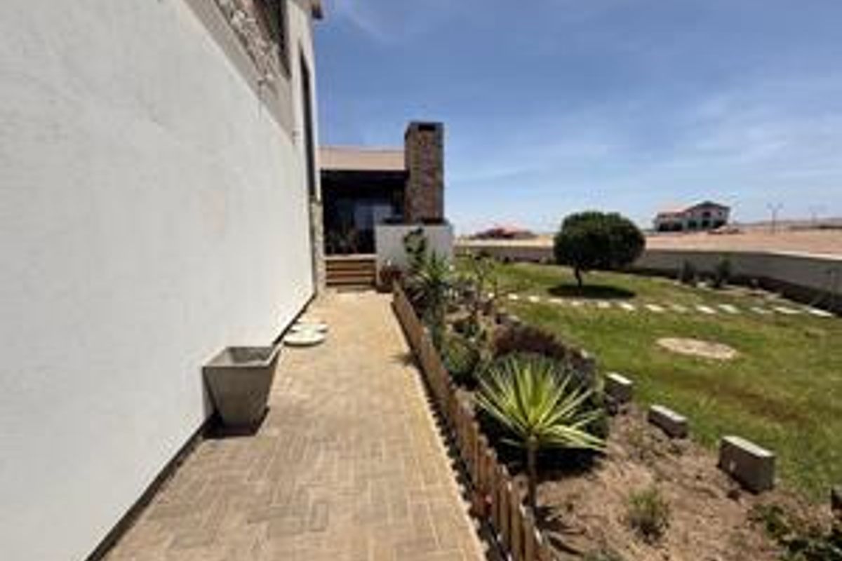 Rear exterior view featuring a paved walkway, stone chimney detail, and landscaped garden area with boundary wall.