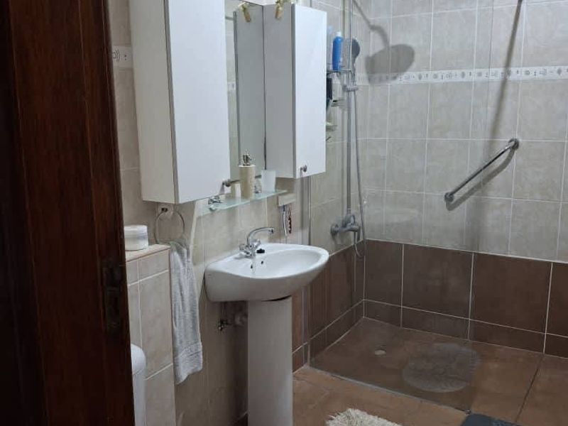 A functional bathroom featuring a walk-in shower with glass screen, pedestal basin, wall-mounted storage cabinet, and neutral floor-to-ceiling tiling.
