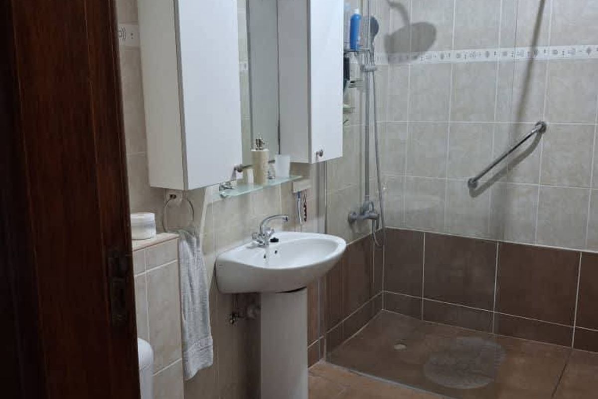 A functional bathroom featuring a walk-in shower with glass screen, pedestal basin, wall-mounted storage cabinet, and neutral floor-to-ceiling tiling.
