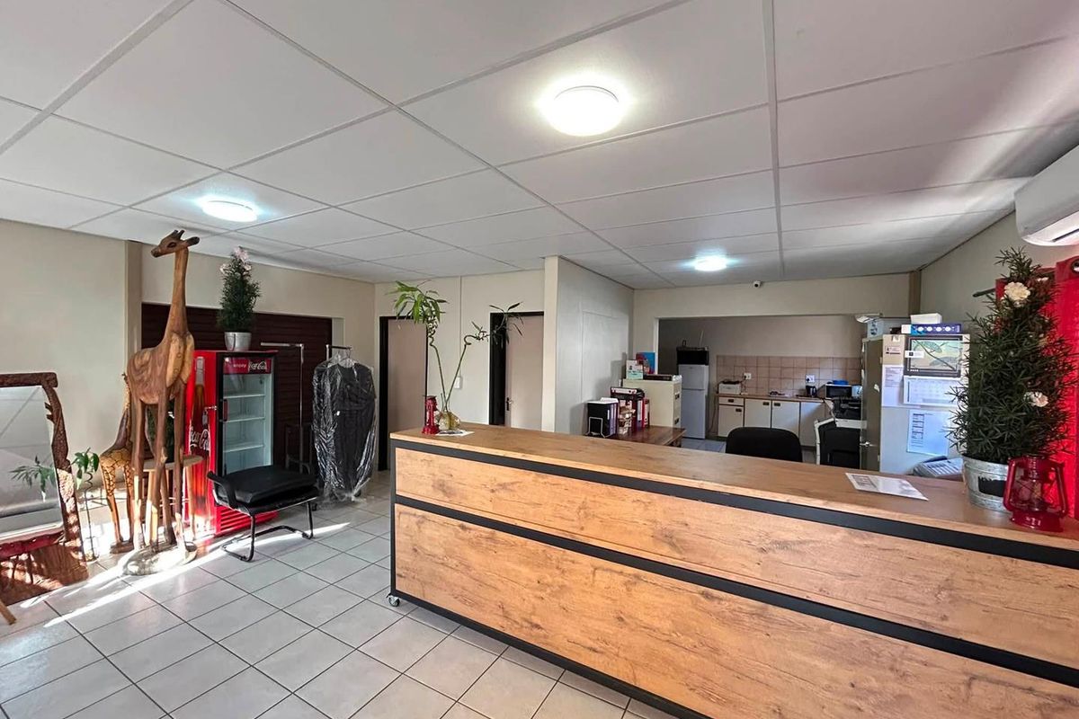 Office reception area featuring a wood-finish desk, tiled flooring, suspended ceiling, and an integrated kitchenette area in the background.
