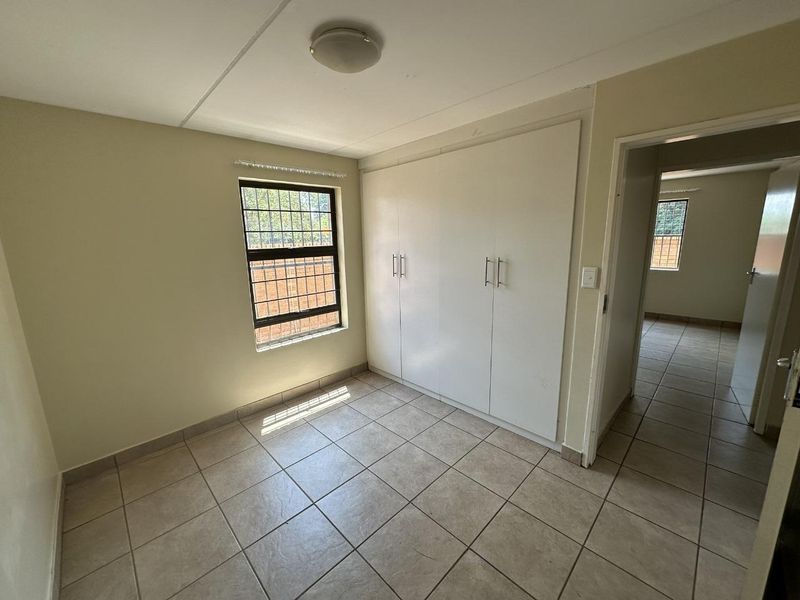A clean, empty bedroom featuring neutral tiled flooring, built-in cupboards, and a window fitted with security bars.