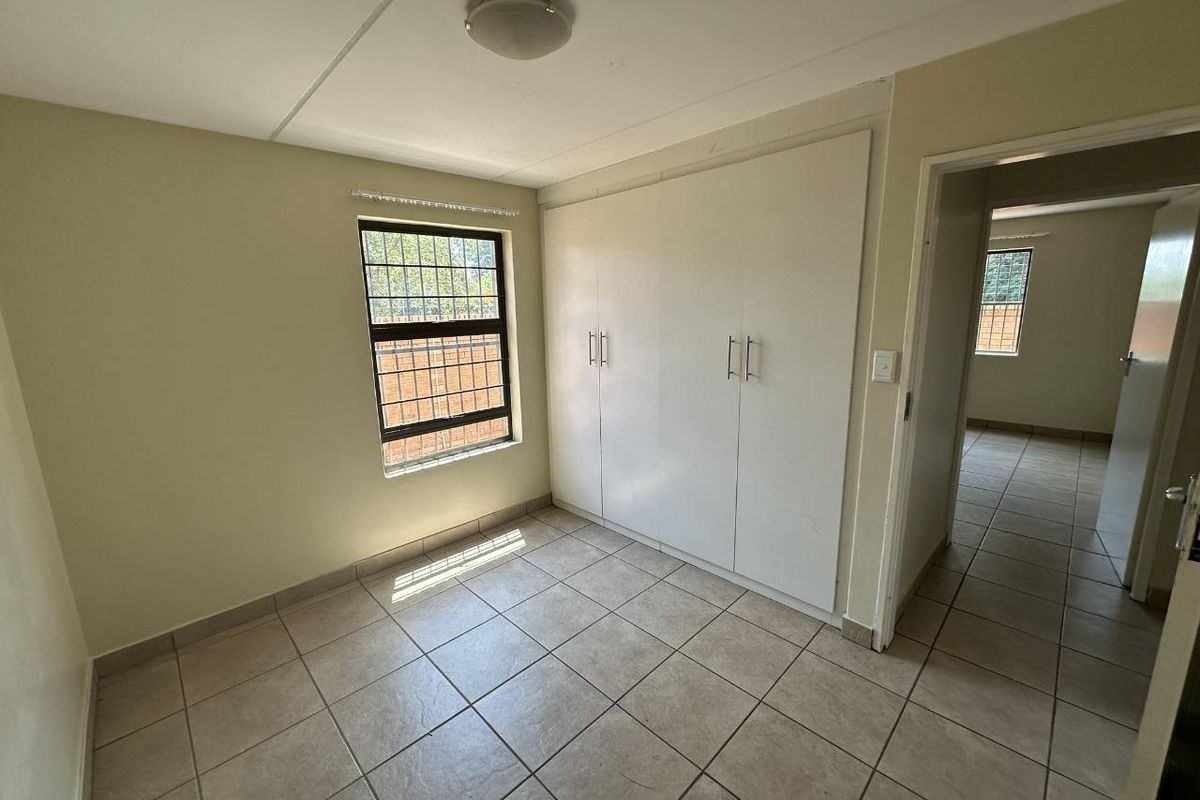 A clean, empty bedroom featuring neutral tiled flooring, built-in cupboards, and a window fitted with security bars.