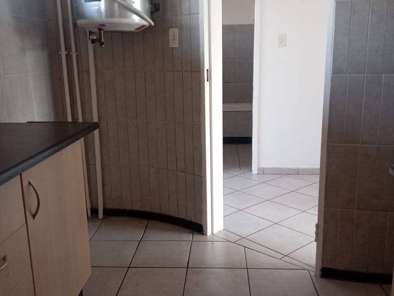 A functional kitchen area featuring tiled flooring, wall splashbacks, built-in cabinetry, and an exposed electric water heater, leading into an adjacent room.