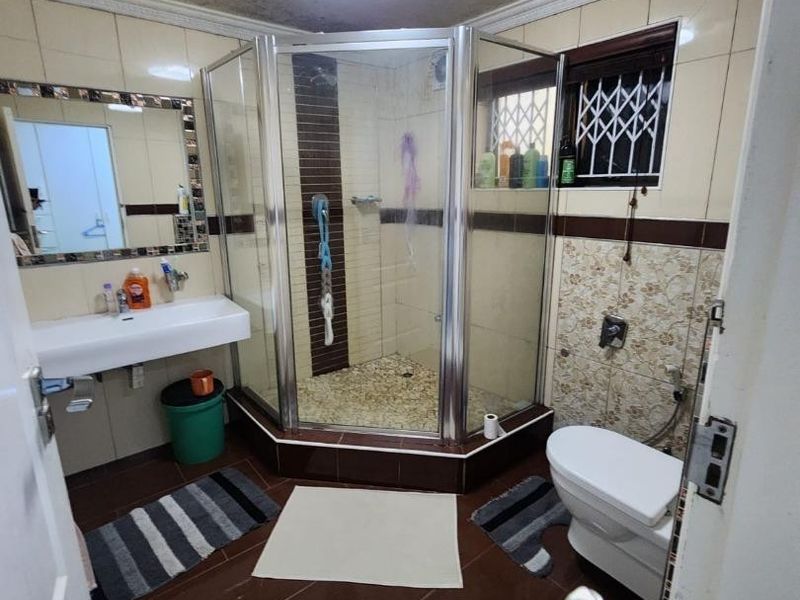 A compact bathroom featuring a corner shower enclosure, wall-mounted basin, and toilet, with tiled finishes throughout and security bars on the window.
