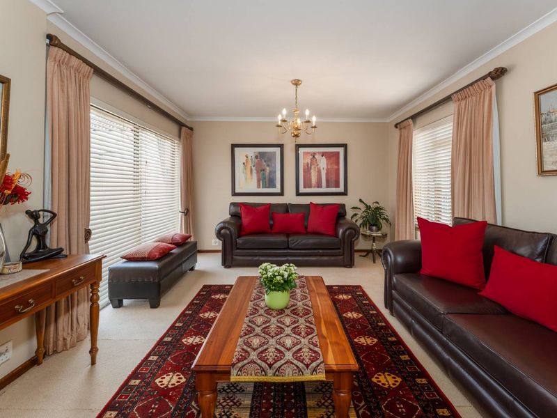 A well-maintained living room featuring neutral walls, carpeted flooring, large windows with venetian blinds, and a central chandelier.