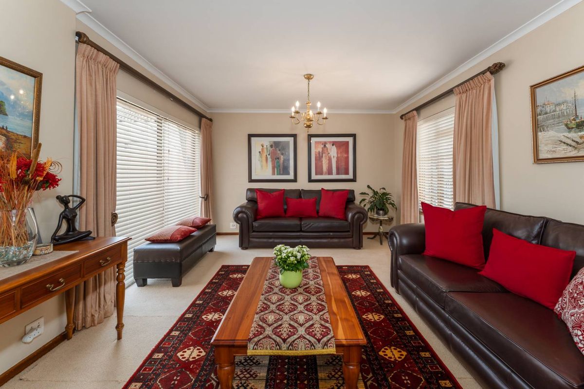 A well-maintained living room featuring neutral walls, carpeted flooring, large windows with venetian blinds, and a central chandelier.