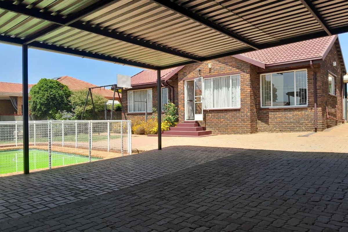 A spacious paved carport area adjacent to a brick-faced house, featuring a swimming pool with safety fencing and a well-maintained garden.
