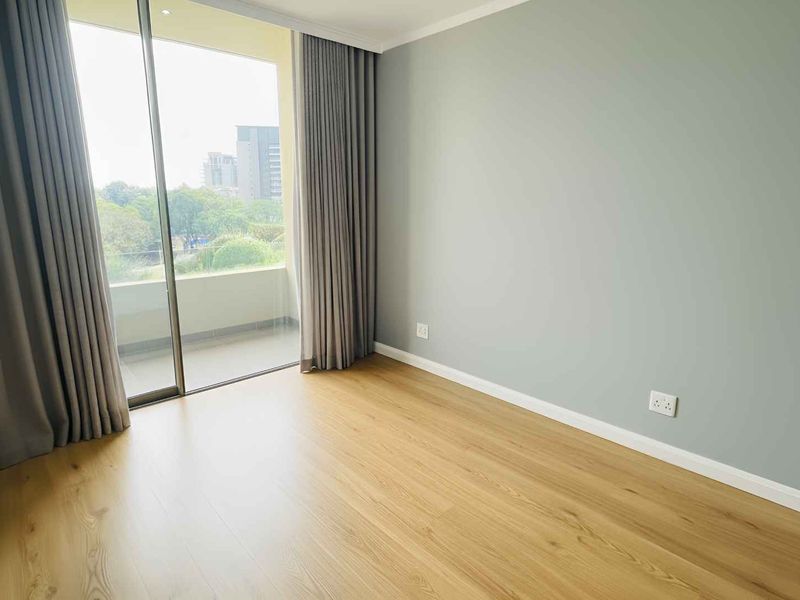 A bright, empty bedroom featuring light wood-look laminate flooring, neutral grey walls, and a large sliding glass door leading to a balcony.