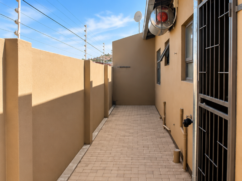 A clean, paved side walkway of a townhouse featuring secure electric fencing, a metal security gate, and functional exterior wall lighting.