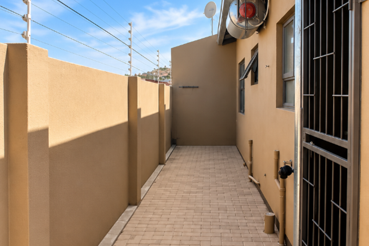 A clean, paved side walkway of a townhouse featuring secure electric fencing, a metal security gate, and functional exterior wall lighting.