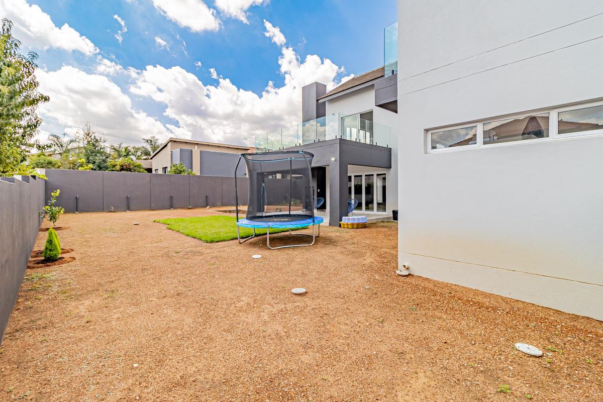 Rear view of a modern double-story home featuring a paved patio, glass balcony railings, and a spacious backyard with perimeter walling.