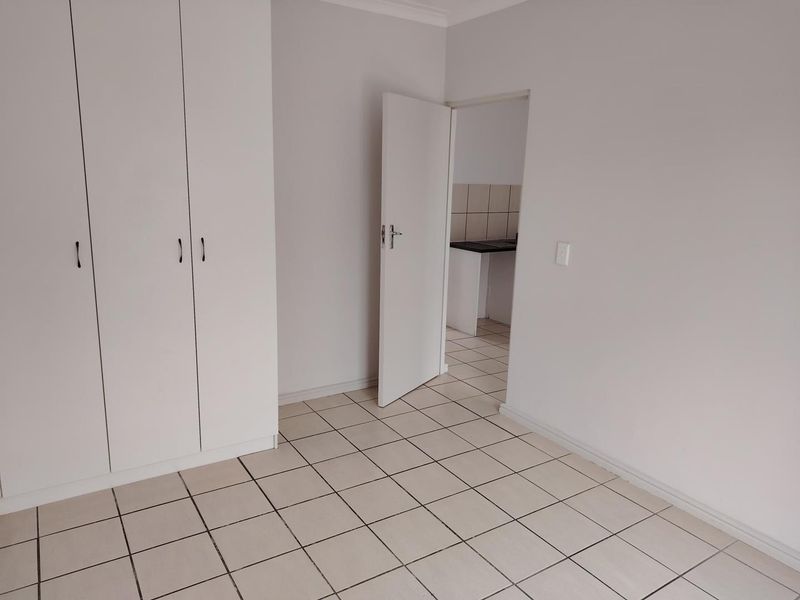 A clean, empty bedroom featuring neutral tiled flooring and white built-in cupboards, with a view into an adjacent kitchen area.