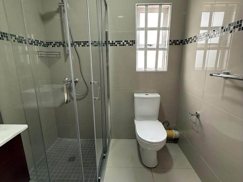 A clean, modern bathroom featuring a glass-enclosed shower, tiled walls and flooring, and a window with security bars.