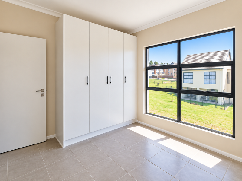 A bright, empty bedroom featuring modern built-in cupboards, neutral tiled flooring, and a large window with a pleasant exterior view.