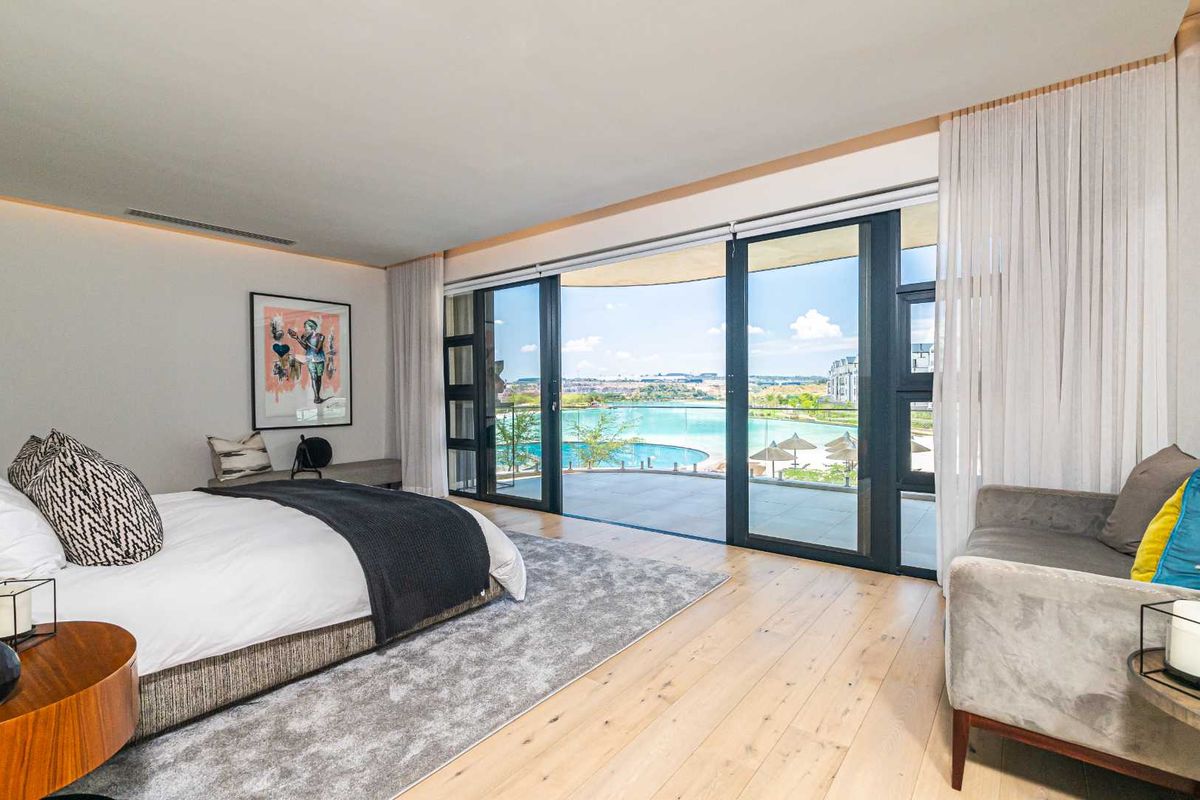 Spacious bedroom featuring light timber flooring, recessed ceiling lighting, and large sliding glass doors opening onto a private balcony with scenic pool views.