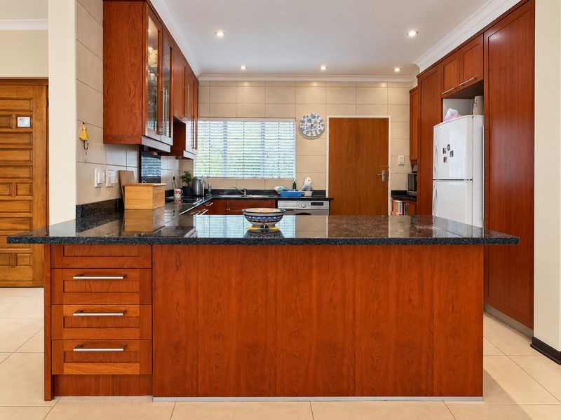 A well-maintained kitchen featuring warm wooden cabinetry, granite countertops, a central island, and neutral tiled flooring in a retirement village setting.