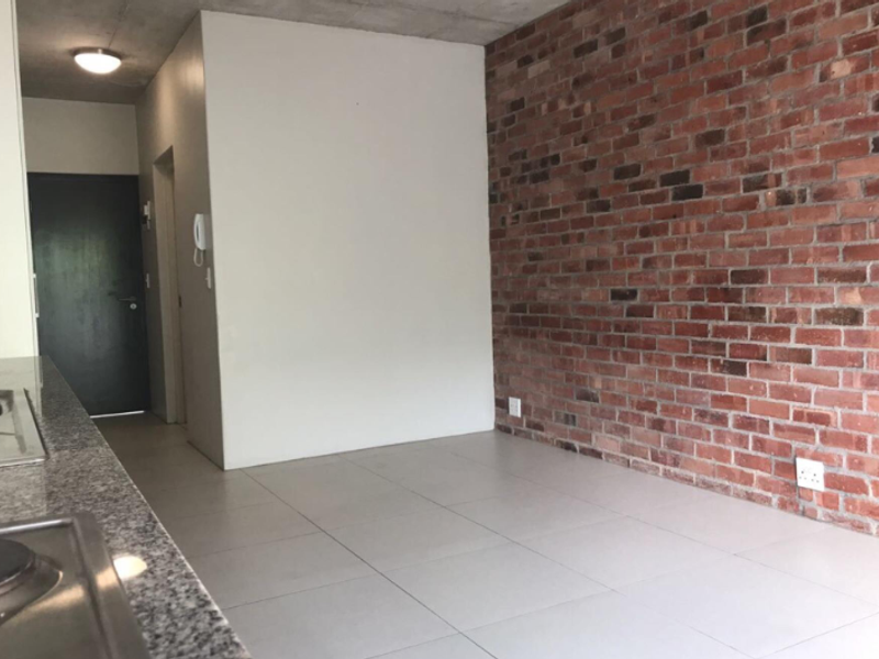 An open-plan living area featuring an exposed brick feature wall, concrete ceiling, tiled flooring, and a modern kitchen counter with a built-in hob.