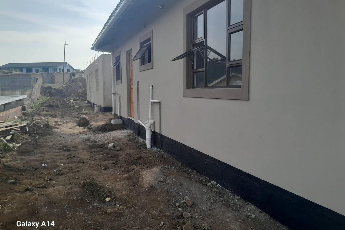Rear exterior view of a house showing plastered walls, aluminium window frames, and exposed plumbing infrastructure with unfinished surrounding ground.