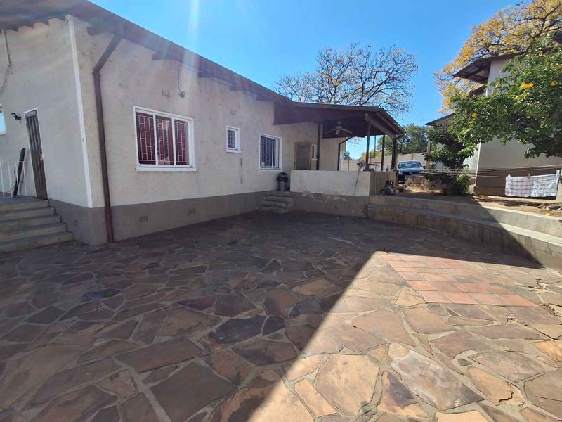 Rear exterior view featuring a stone-paved patio area, a covered veranda, and established garden surroundings with security-fitted windows.