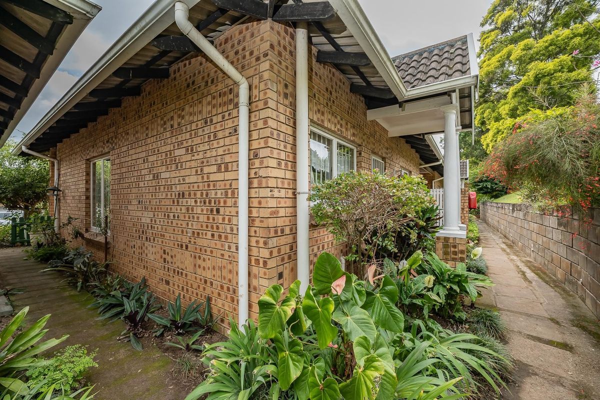 Side view of a face brick townhouse featuring a tiled roof, exposed rafters, and a paved walkway surrounded by established garden beds.