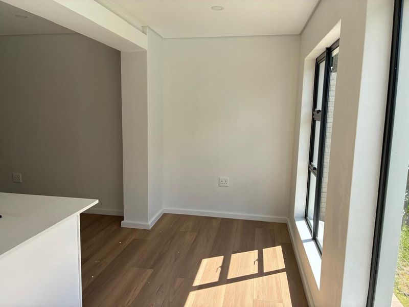 A bright, empty living area featuring modern laminate flooring, clean white walls, and large black aluminium windows allowing for ample natural light.