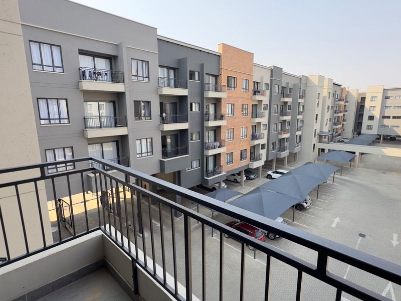 Elevated view of an apartment complex exterior featuring a balcony, paved parking area with shade structures, and modern multi-story building facades.