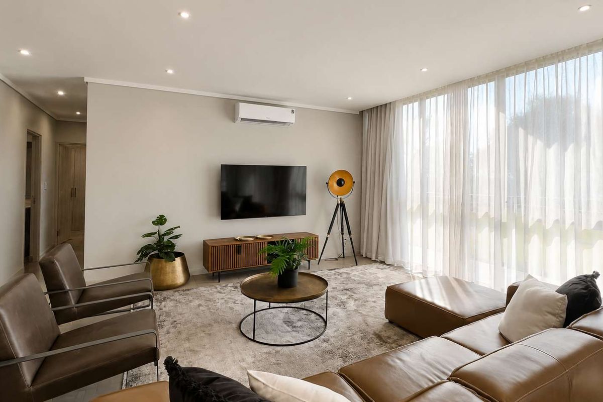 A bright and modern living room featuring recessed downlights, a wall-mounted air conditioning unit, and large floor-to-ceiling windows with sheer curtains.