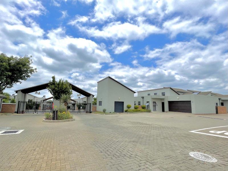 A wide-angle view of the townhouse complex entrance, featuring a paved driveway, secure gated access, and modern residential building facades.
