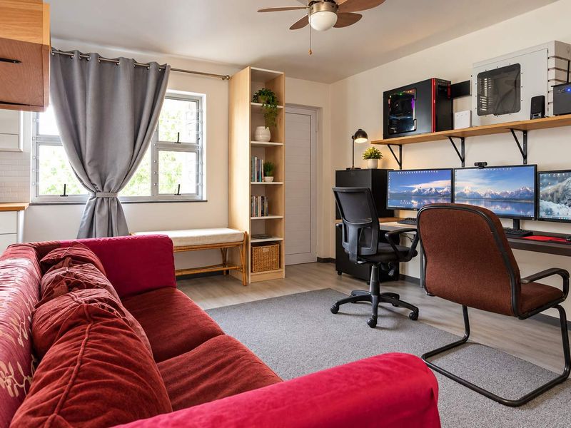 An open-plan living area featuring laminate flooring, a ceiling fan, and a window, with a functional layout that integrates a workspace.
