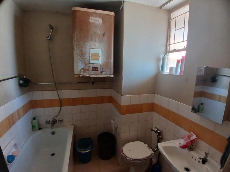 A dated bathroom featuring a bathtub, pedestal basin, toilet, and a wall-mounted electric water heater showing significant signs of corrosion and wear.