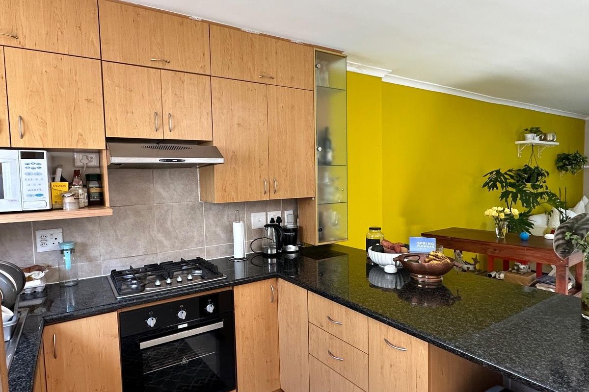 A functional kitchen featuring light wood cabinetry, granite countertops, a gas hob, and an integrated oven, opening into a bright living area.