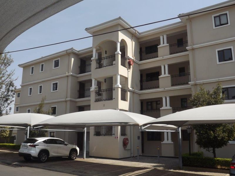 Exterior view of a multi-story apartment building featuring balconies, covered parking bays, and paved driveway access.